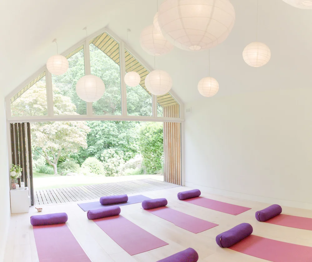 Wide interior view of the yoga studio showing a vaulted white ceiling filled with many spherical paper lanterns. A large gable-shaped window and folding doors open to a sunlit garden and a wooden deck, with trees and greenery visible outside. Inside, multiple yoga mats in pink and purple are neatly laid out in two rows, each with a purple bolster at the top, arranged for a class. The space is bright and minimal, with pale wood floors and white walls.