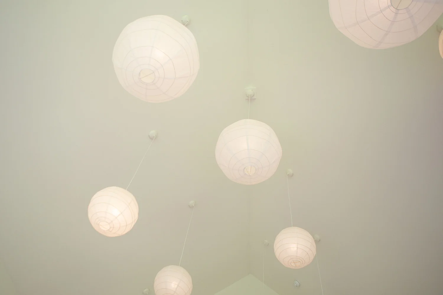 Close-up looking up at the studio ceiling, showing several white spherical paper lantern lights suspended by thin cords at different heights. The ceiling is smooth and pale, and the lanterns glow softly, emphasizing a calm, airy atmosphere. Only the ceiling and lights are visible, with no walls or floor in frame.