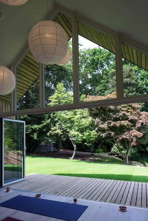 Interior view looking out through the open gable window and folding door toward a lush garden. A large white paper lantern hangs in the upper left, and the wooden deck is visible below. A single blue yoga mat lies near the threshold with a few small tea-light candles placed along the edge, creating a tranquil, atmospheric scene.
